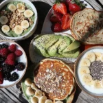 A beautiful gluten-free lactose-free breakfast spread with pancakes topped with fresh berries and maple syrup, coconut yogurt with bananas and chia seeds, avocado toast on gluten-free bread with hemp seeds, a spinach smoothie bowl with granola, and scrambled eggs made with dairy-free milk, arranged on a rustic wooden table in natural sunlight.